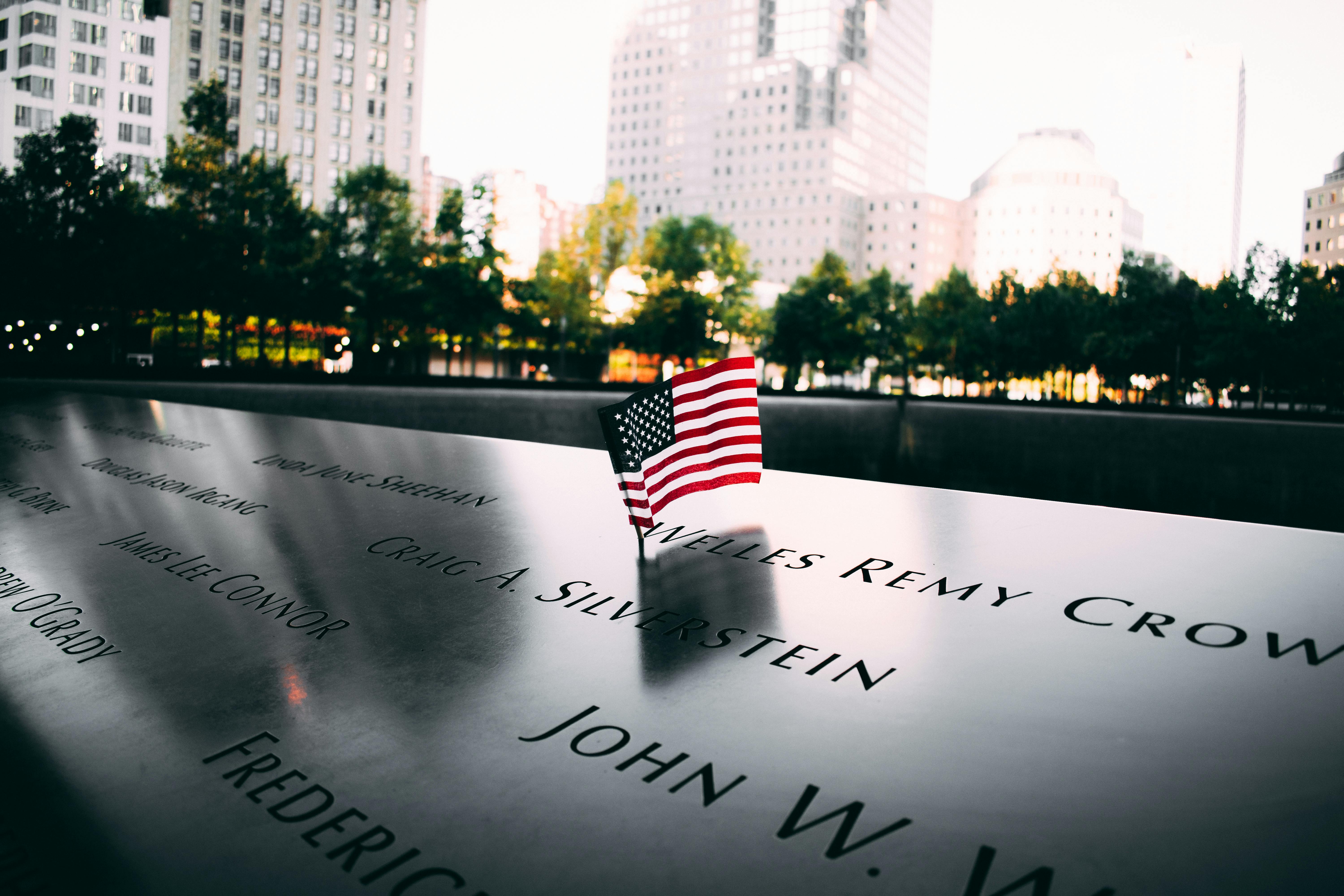 9/11 Memorial pool with engraved names, small American flag, and NYC skyline in background.